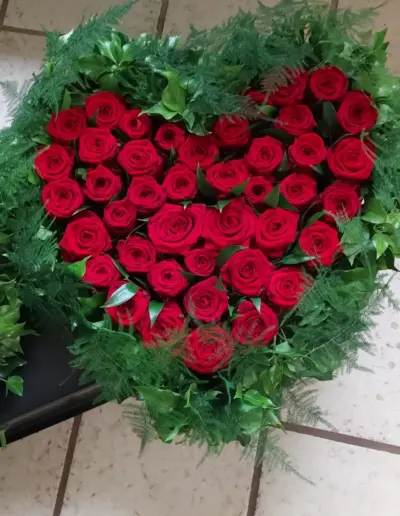 Herzförmiges Trauergesteck aus roten Rosen, umrahmt von dichtem grünem Beiwerk.