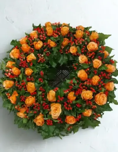 Trauerkranz mit orangefarbenen Rosen, roten Beeren und grünem Blattschmuck.