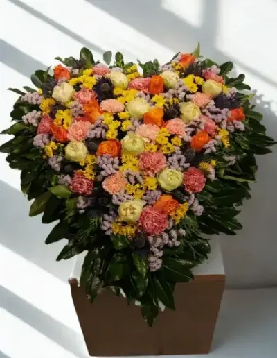 Herzförmiges Trauergesteck mit Rosen in Orange und Creme, Nelken und bunten Blüten auf grünem Blattschmuck.
